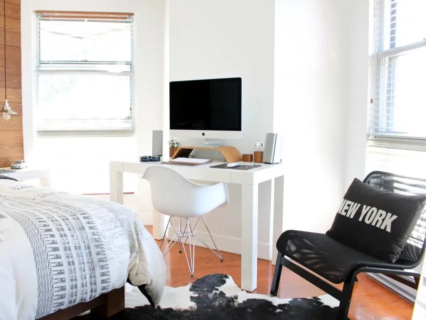 A bedroom painted in a flat white, with a made bed, a desk under the window and bare floorboards.