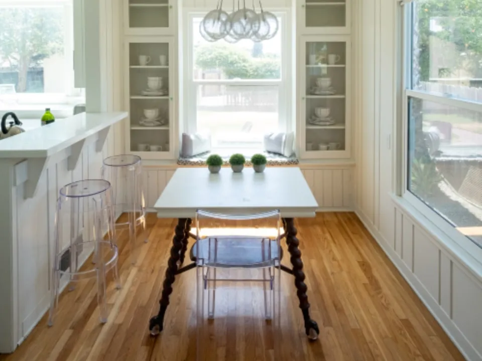 A breakfast nook with built-in glass-fronted cabinetry painted off-white, lit by a tall bay window.