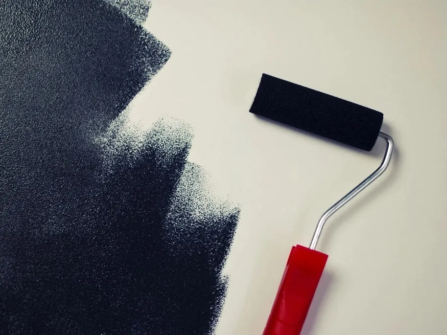 A paint roller with a red handle lying on a cream wall beside a rough-edged band of near-black paint, part way through a first coat.