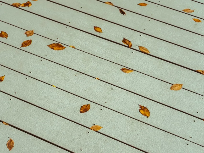 Deck boards finished in a soft sage solid stain, with a few fallen autumn leaves scattered across them.