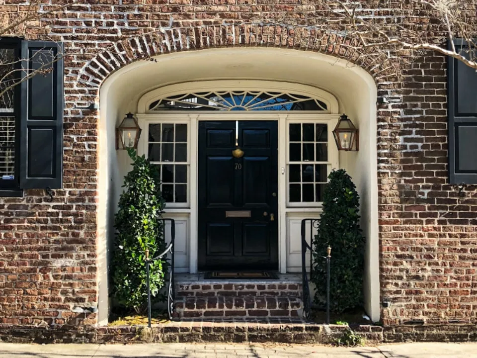 A brick house with a white arched portico, black front door, black shutters and carriage lamps either side.