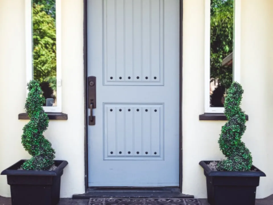 A panelled front door painted soft blue-grey, framed by crisp white trim and a pair of clipped topiaries.